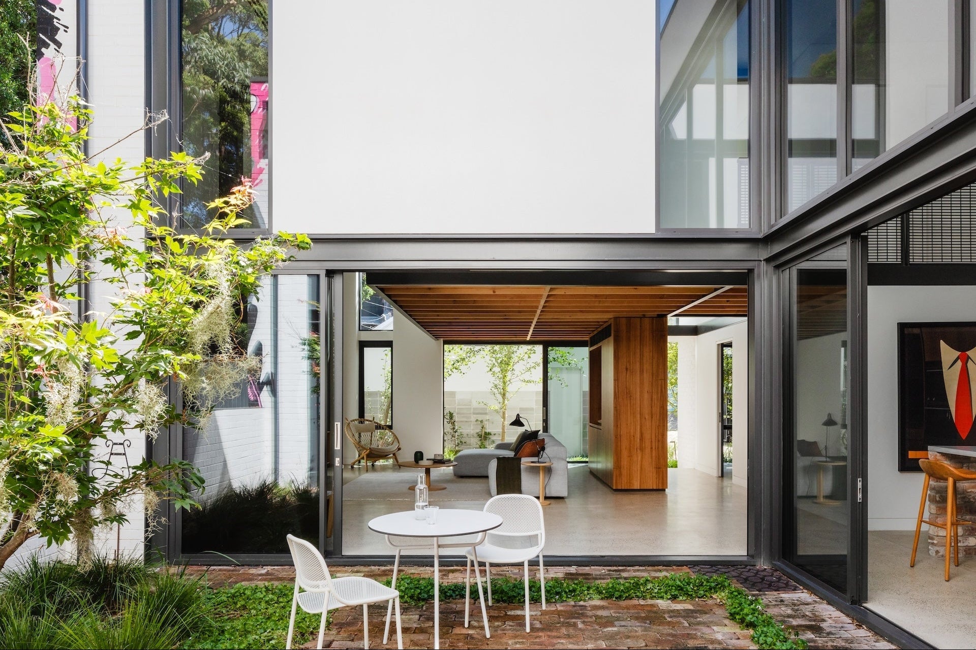 Modern two-story house exterior with large black-framed glass windows, white walls, a small outdoor patio table and chairs, and visible interior living space through the open lower level.